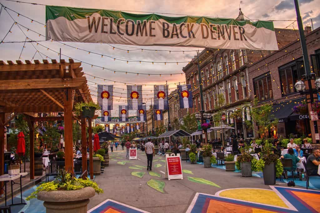 A painted pedestrian street with stores on either side, string lights across the middle, Colorado flags over the street, and a large size that reads "Welcome back Denver".