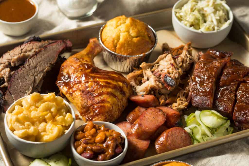A platter with bbq chicken, sausage, brisket, ribs, baked beans, mac and cheese, mashed potatoes, and corn bread.