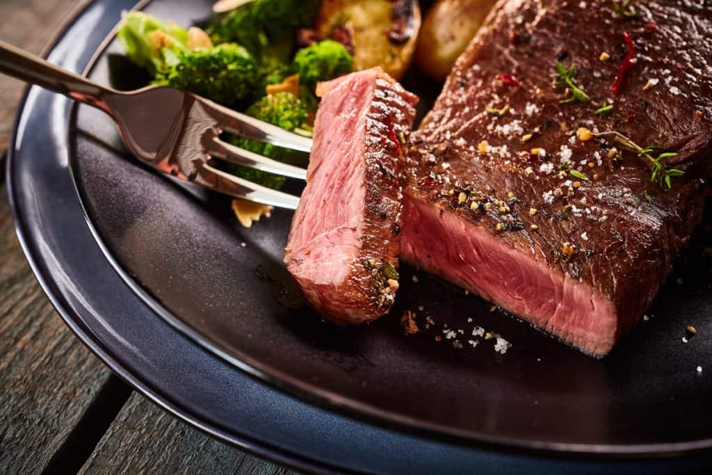 A fork with a slice of steak help above a plate with steak and broccoli. 