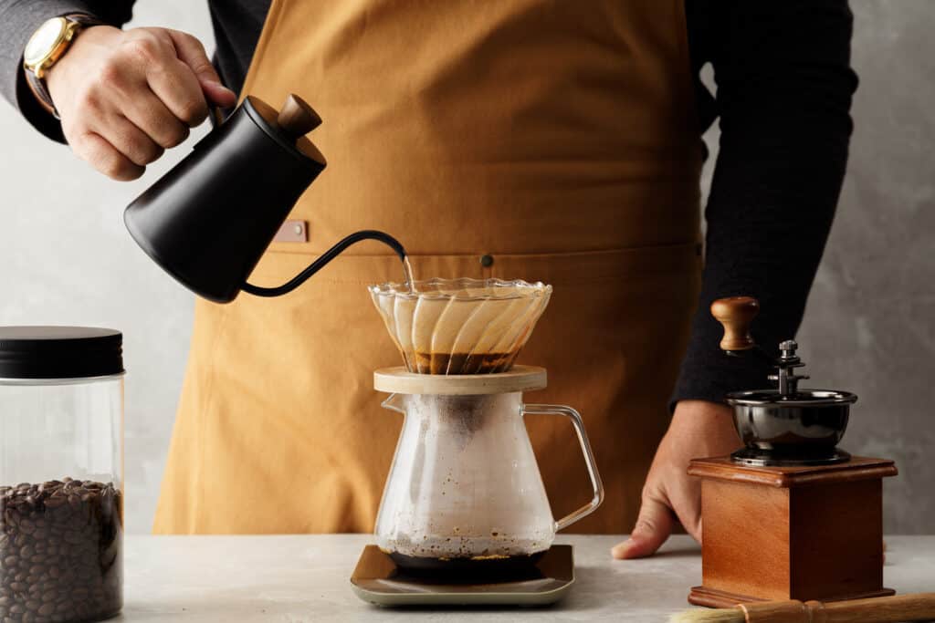 Pour-over coffee brewing process with hot water being poured from a black kettle into a filter over a carafe, with a manual coffee grinder on the side, embodying the artisanal coffee-making ritual.