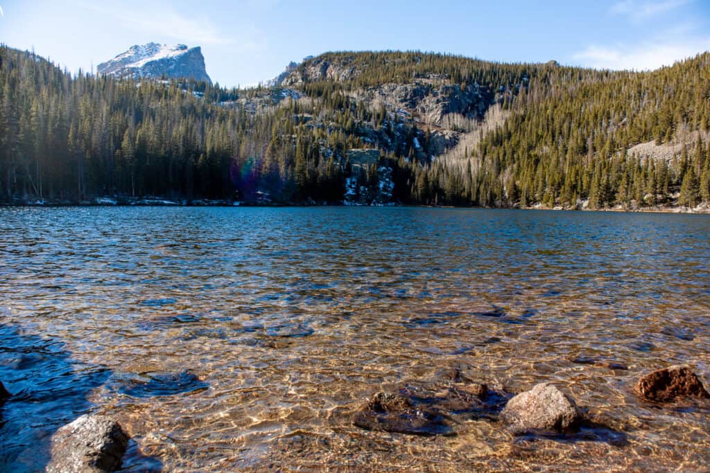 The crisp waters of Bear Lake in Rocky Mountain National Park ripple gently near the shore, where rocks are visible through the clear water. Towering pine trees and rugged mountain slopes with patches of snow under a bright blue sky frame the serene lake.