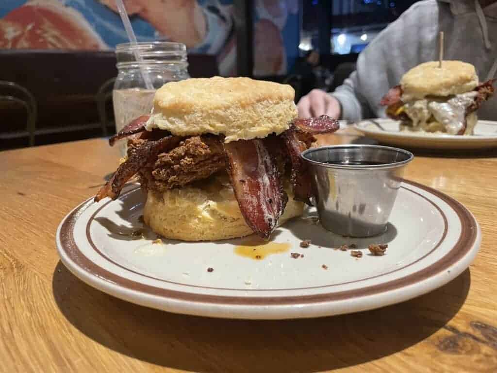 A biscuit sandwich with crispy fried chicken and bacon strips served on a plate with a side of syrup, in a casual dining setting.