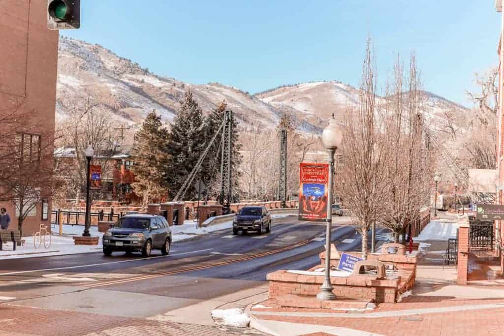 A calm street scene in downtown Golden, Colorado, with traffic lights, bare winter trees, and vehicles passing by. The city's architecture and the Clear Creek History Park's bridge frame the snowy foothills of the Rocky Mountains in the distance, showcasing the town's blend of historic charm and natural beauty.