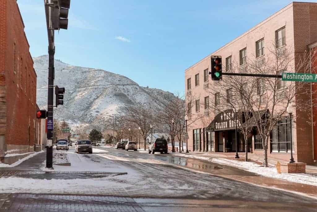 A quiet winter day in Golden, Colorado, with the Golden Hotel facing the street corner under a traffic light on Washington Avenue, with the majestic snow-dusted Table Mountain in the background, creating a postcard-like setting.