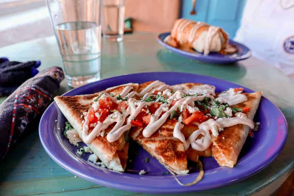 A mouth-watering quesadilla quartered on a purple plate, garnished with fresh diced tomatoes, a generous drizzle of sour cream, and chopped herbs. Served on a teal table with a casual setting, accompanied by a glass of water and another burrito dish in soft focus in the background, ready for a delicious meal.