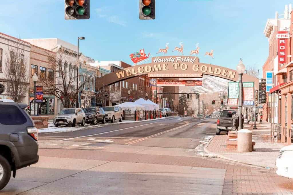 The iconic "Welcome to Golden" archway greets visitors with a "Howdy Folks!" in vibrant red and gold, framing the view of the historic downtown area of Golden, Colorado, where the West lives, complete with a backdrop of a clear sky and a hint of the Rocky Mountains.