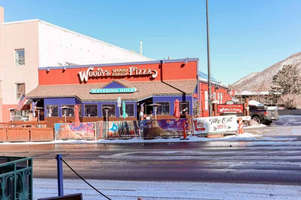 The vibrant red facade of Woody's Wood Fired Pizza in Golden, Colorado, stands out on a sunny day, with American flags flying above the "Watering Hole" sign and the snowy street and mountain backdrop inviting patrons in for a cozy dining experience.